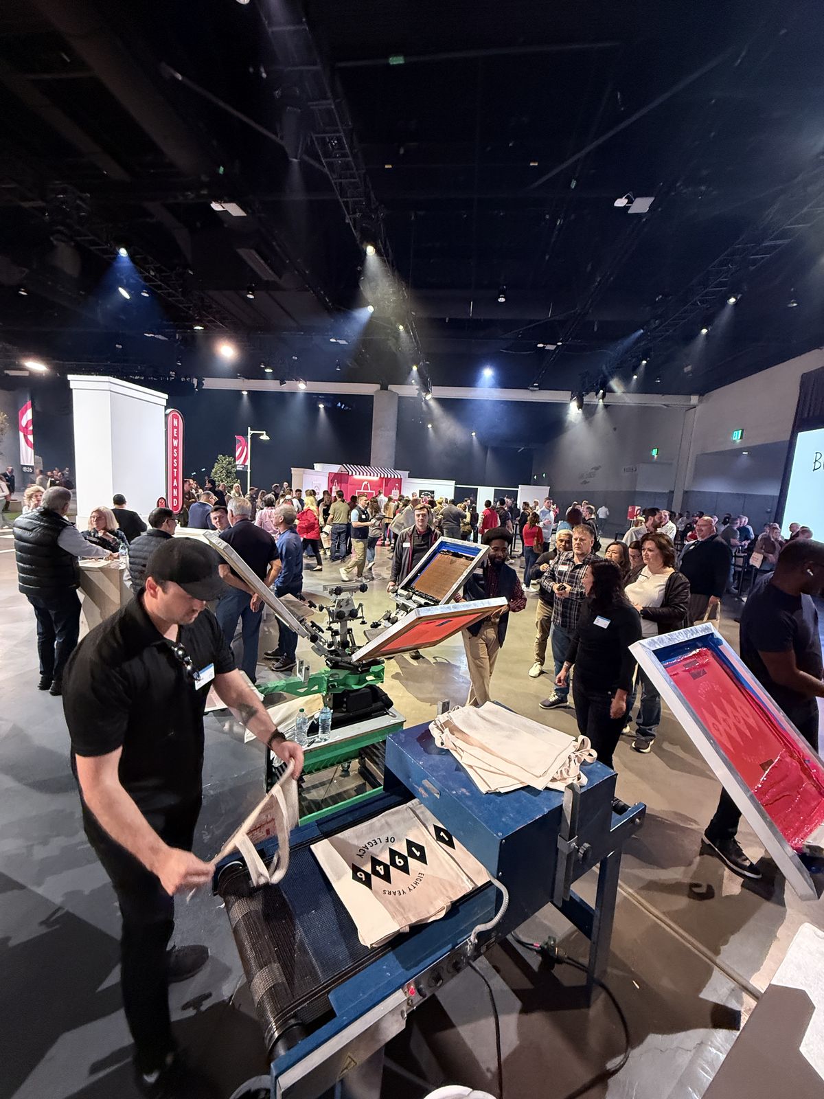 Wide view of a Merch Troop station with a conveyor dryer and anniversary totes inside a convention hall